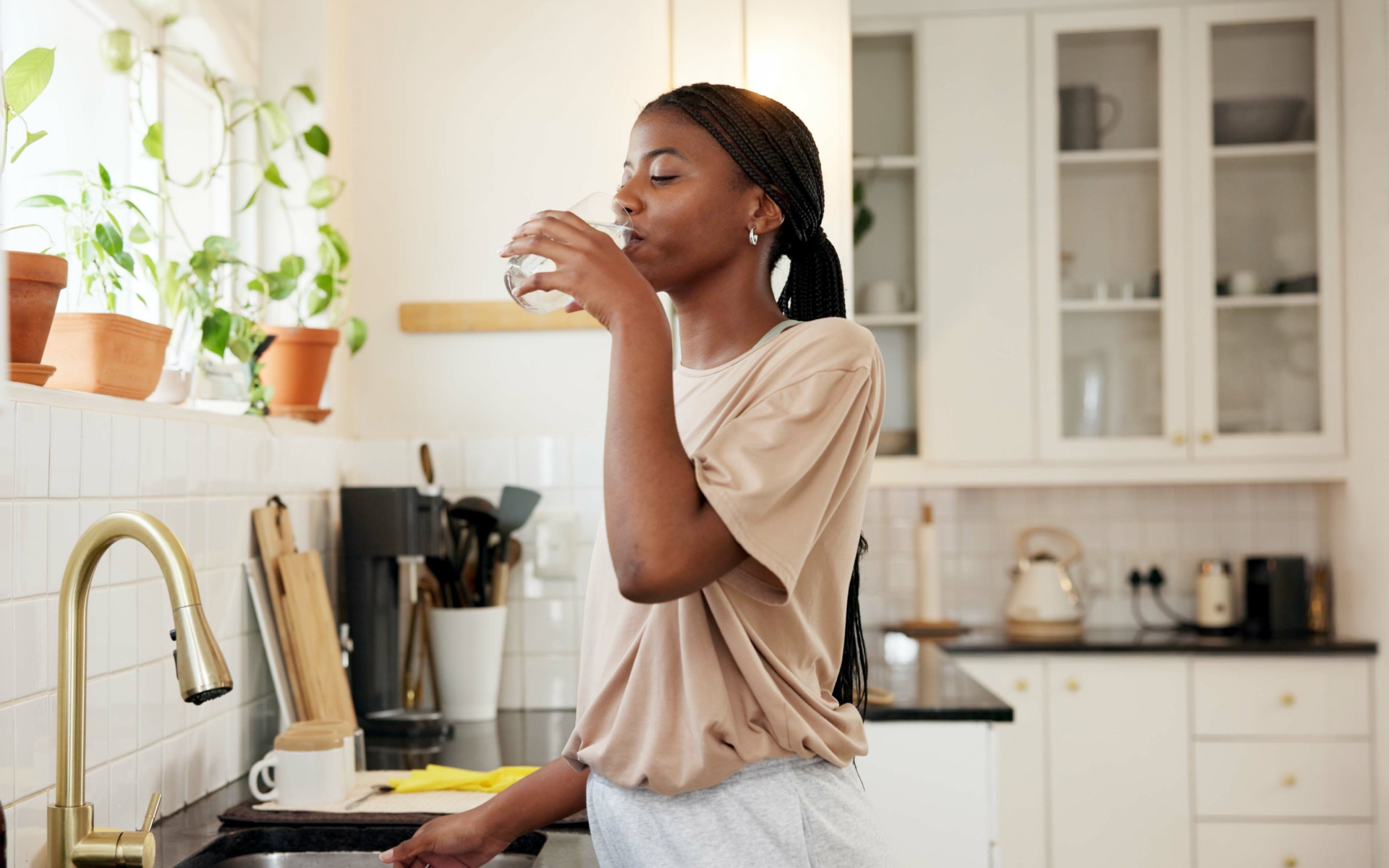 Woman drinking water in a kitchen, illustrating how much water should I drink while fasting and healthy hydration habits.