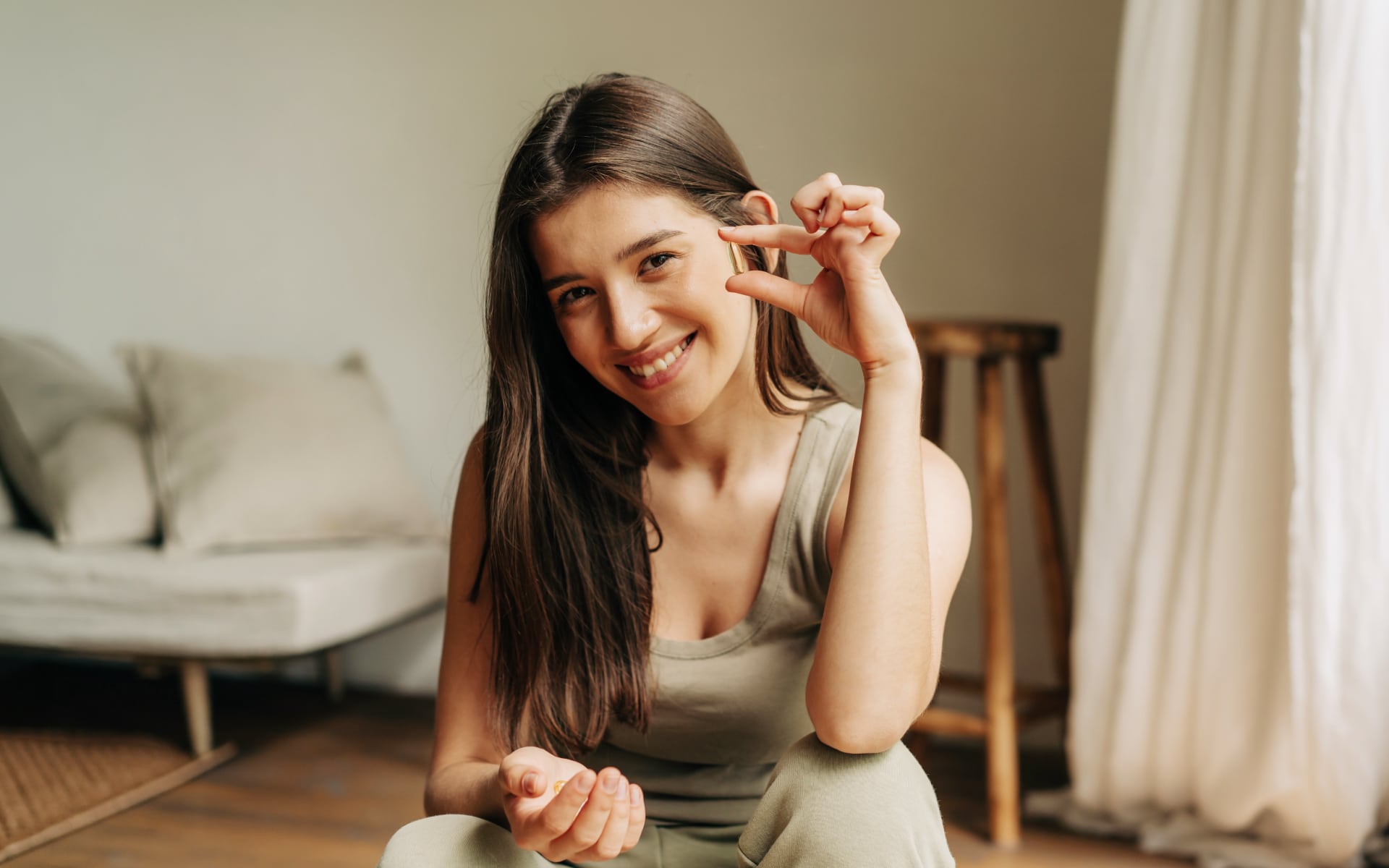 Smiling woman holding a vitamin capsule between her fingers at home, illustrating the question, “Do vitamins break a fast?”