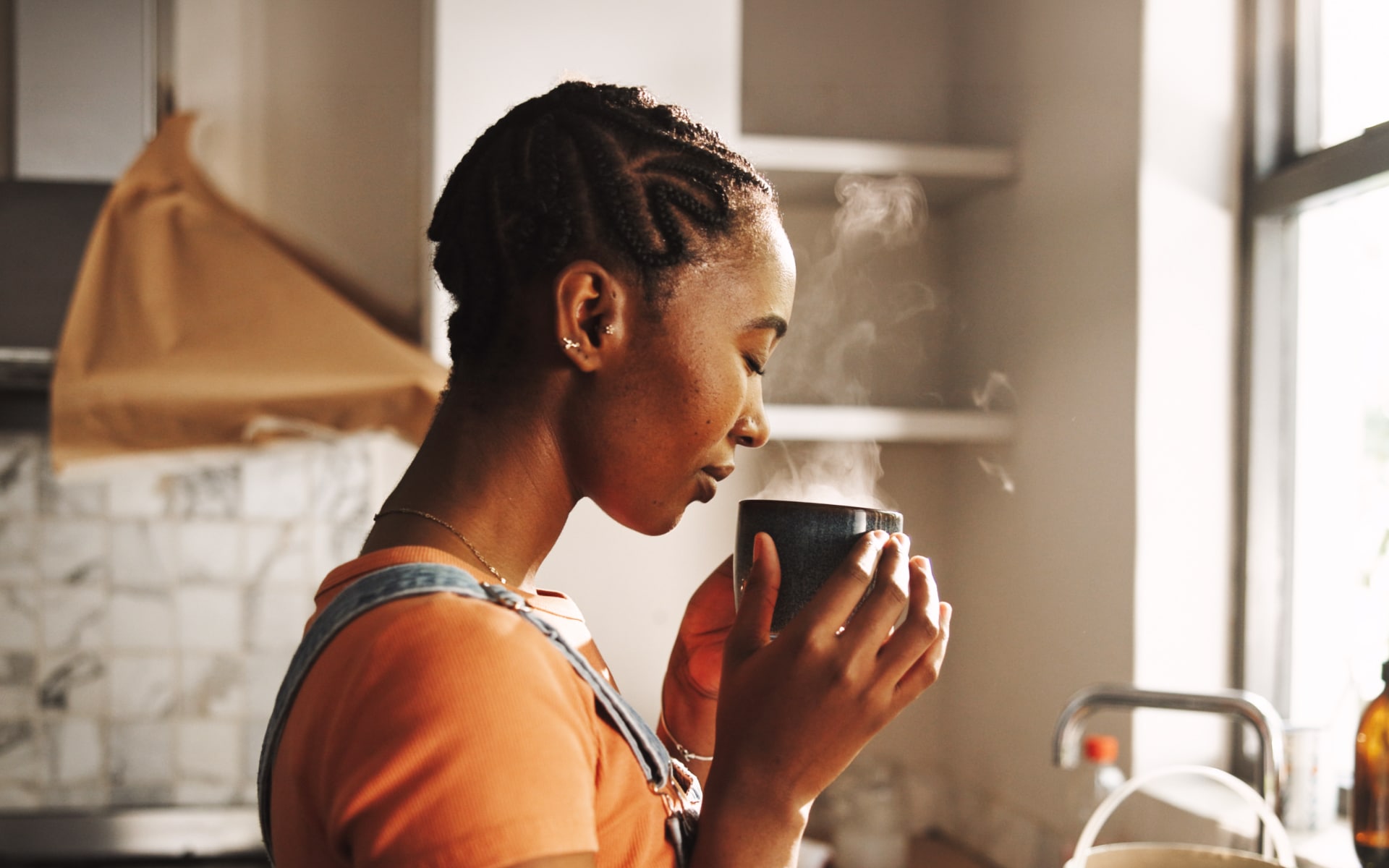 Woman drinking a steaming cup of coffee in a sunlit kitchen, illustrating the question, “Does coffee break a fast?”