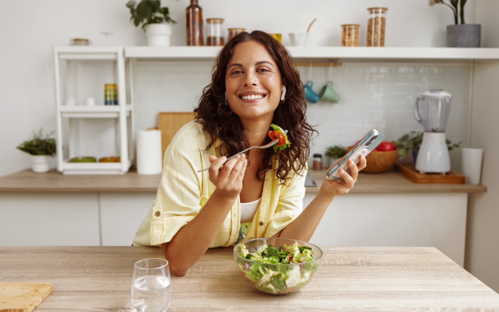 Woman smiling at a kitchen table with a bowl of salad and phone in hand, illustrating a low carb intermittent fasting routine in a bright home setting.