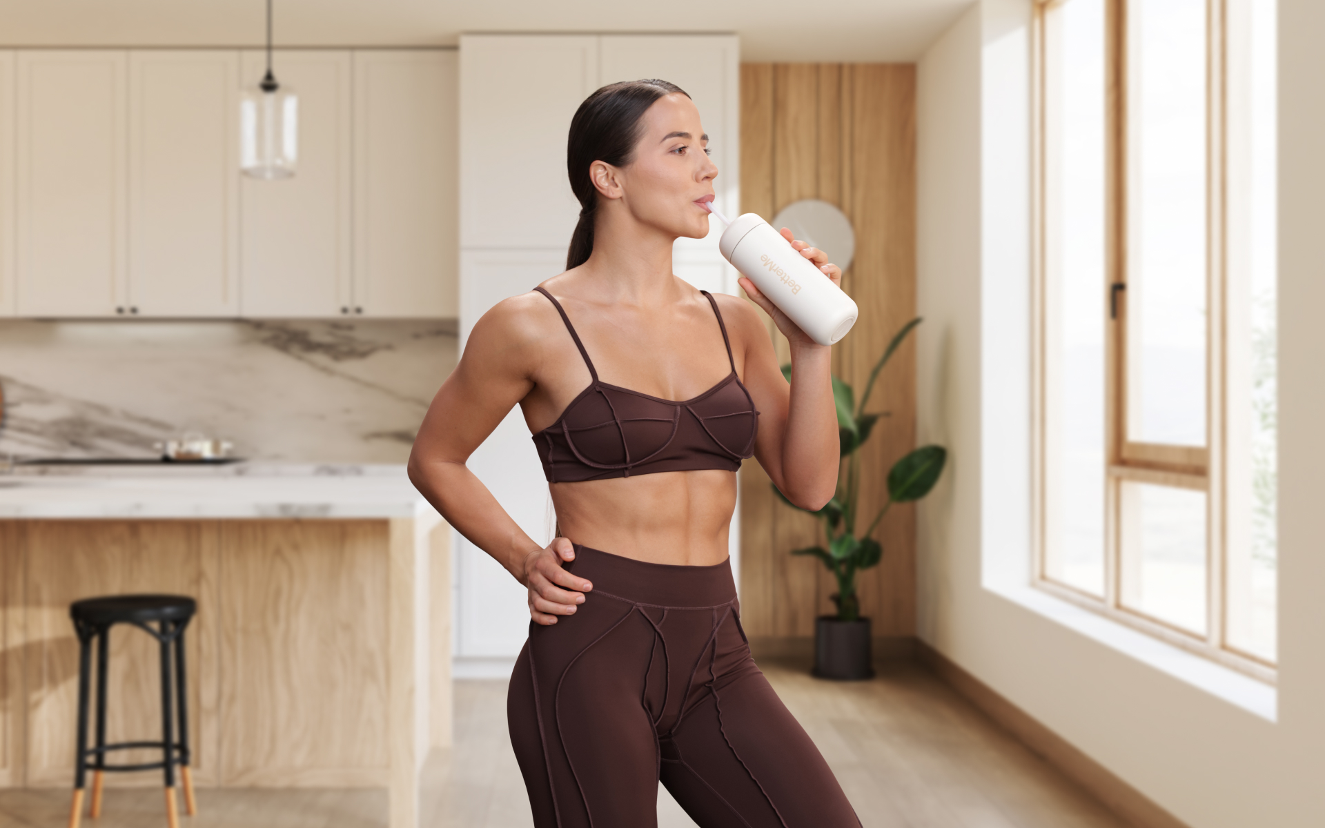 Woman in workout clothes drinking from a bottle in a bright kitchen, representing hydration support during a 5 day water fast.