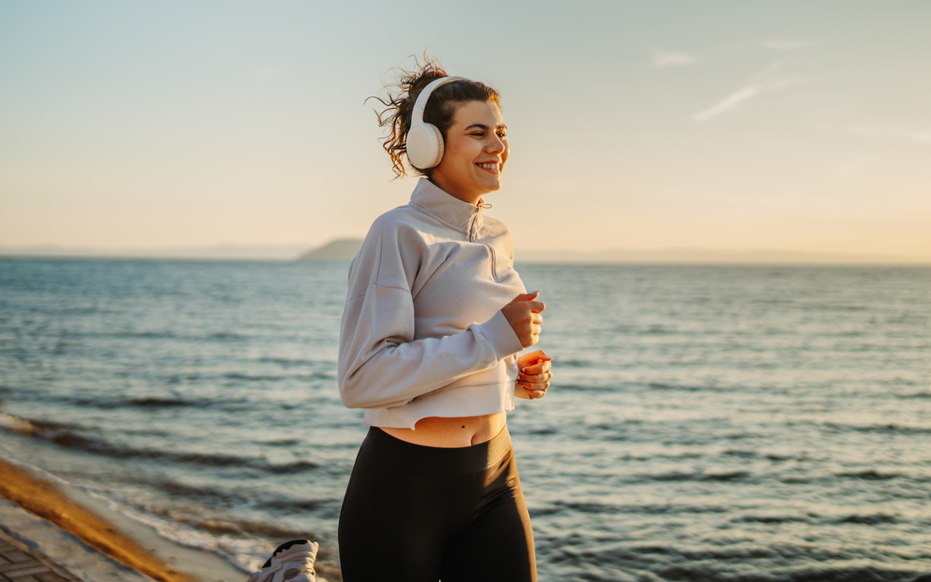 Woman jogging by the sea at sunset while wearing headphones, illustrating a daily routine to run 5 miles a day.