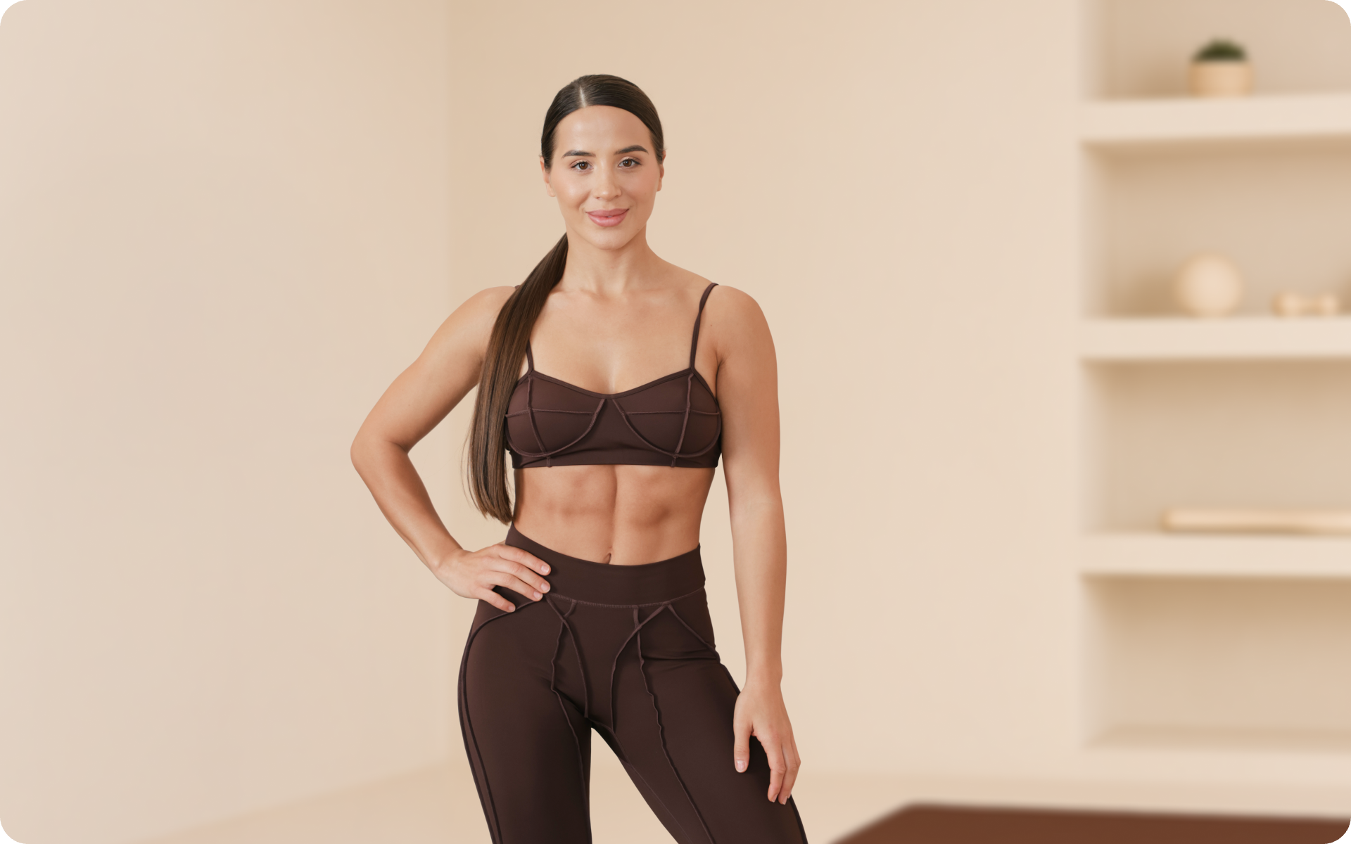Lessie Fitness stands in a chocolate brown activewear set in a bright, neutral studio, posing with one hand on her hip.