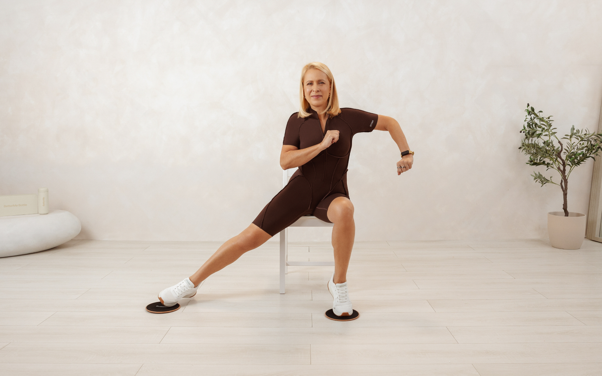 Woman demonstrating seated leg exercises for seniors, sitting on a chair with one leg extended to the side in a bright home workout space.