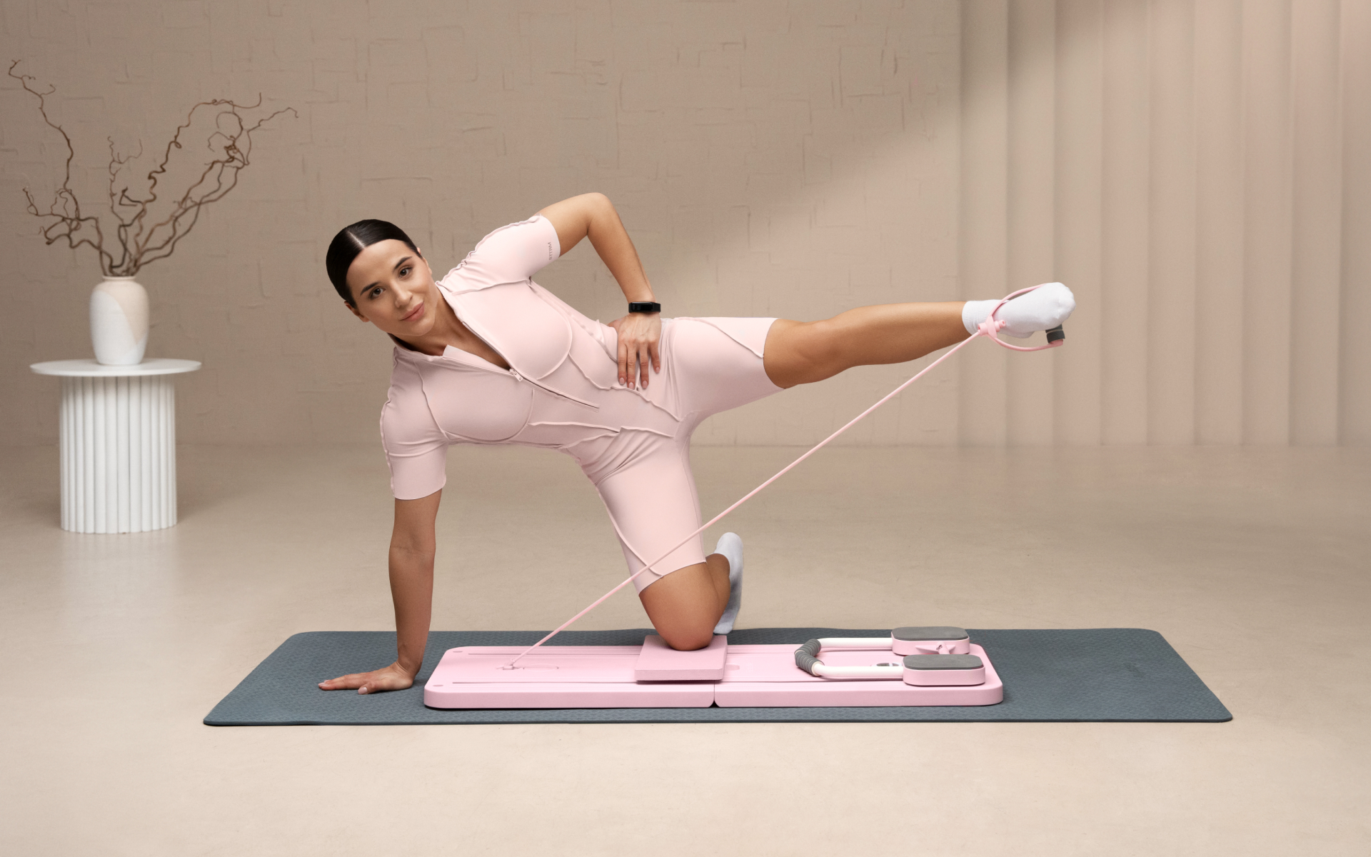 Woman demonstrating one of the advanced pilates reformer exercises on a compact pink mini reformer, balancing on one knee with one leg extended against resistance in a bright studio.