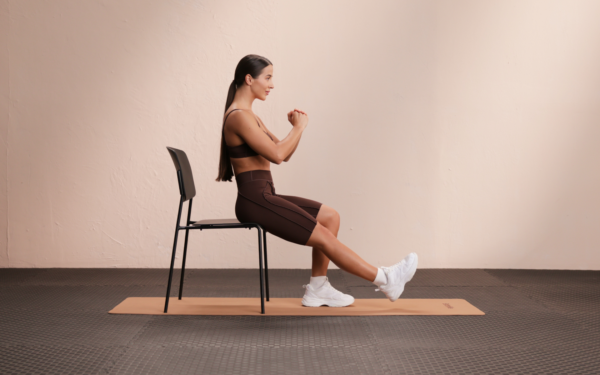 Woman demonstrating a seated forward fold stretch on a chair, sitting tall with one leg extended forward and hands held in front of her chest on a workout mat.