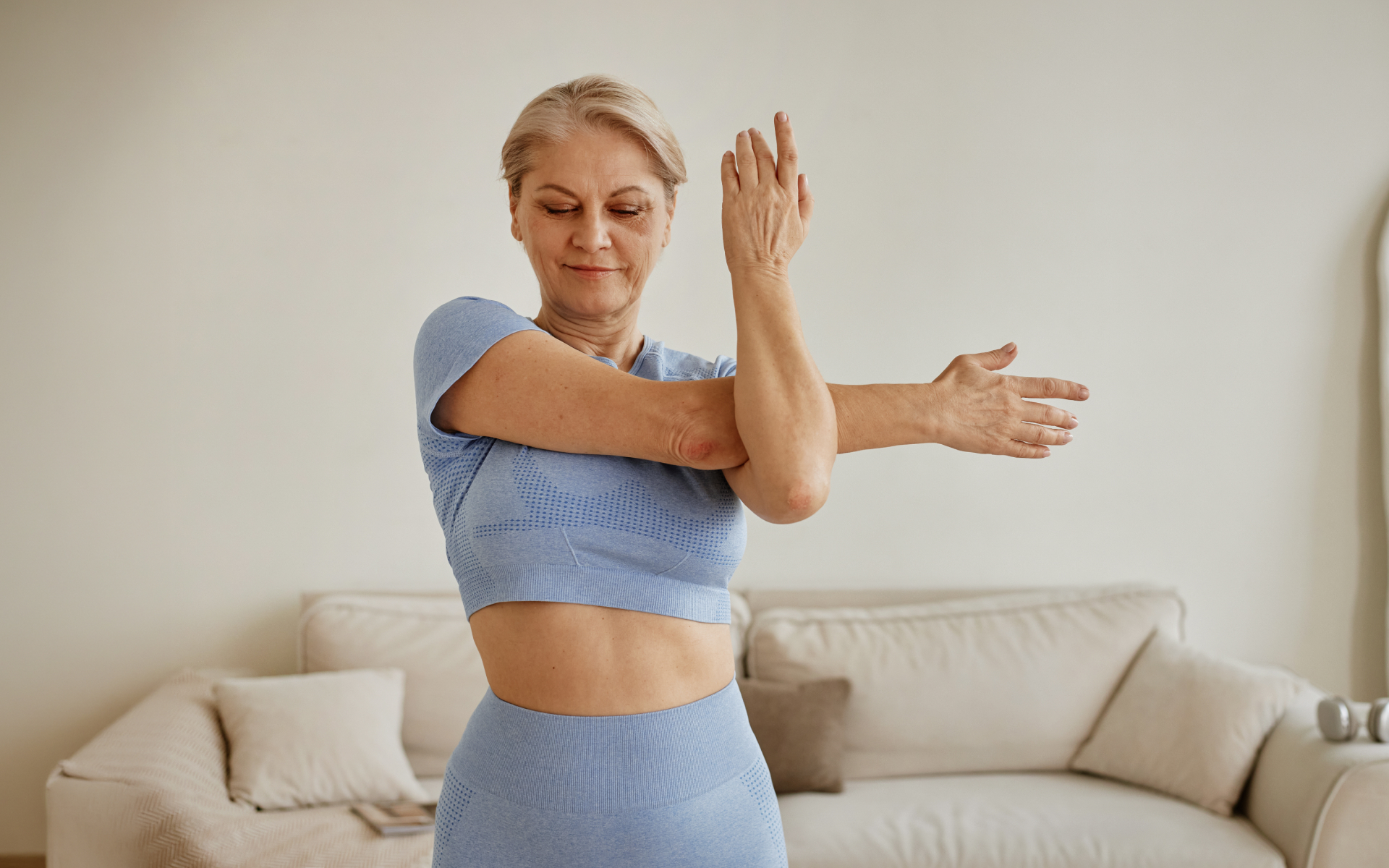 Older woman stretching her shoulders in a bright living room, demonstrating one of 10 flexibility exercises for mobility and upper-body flexibility.