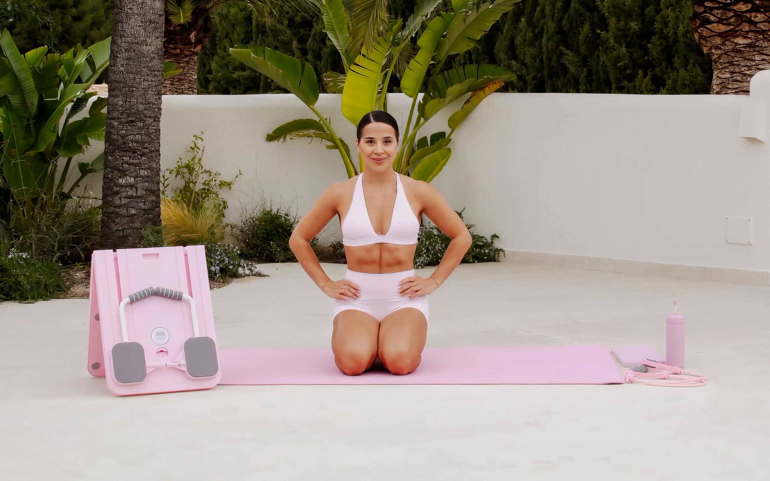 Lessie Fitness kneeling on a pink mat outdoors, wearing a light pink workout set, with a BetterMe mini reformer and accessories placed beside her.