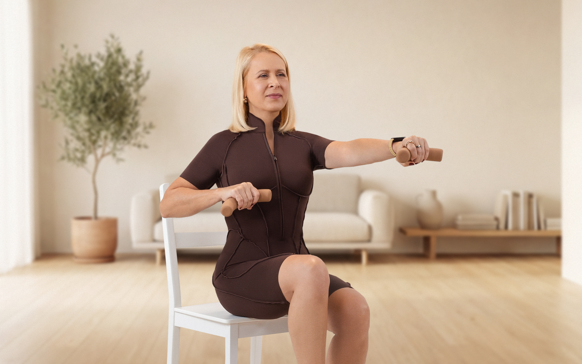 Woman performing seated arm exercises for seniors with light hand weights while sitting on a chair in a bright room.