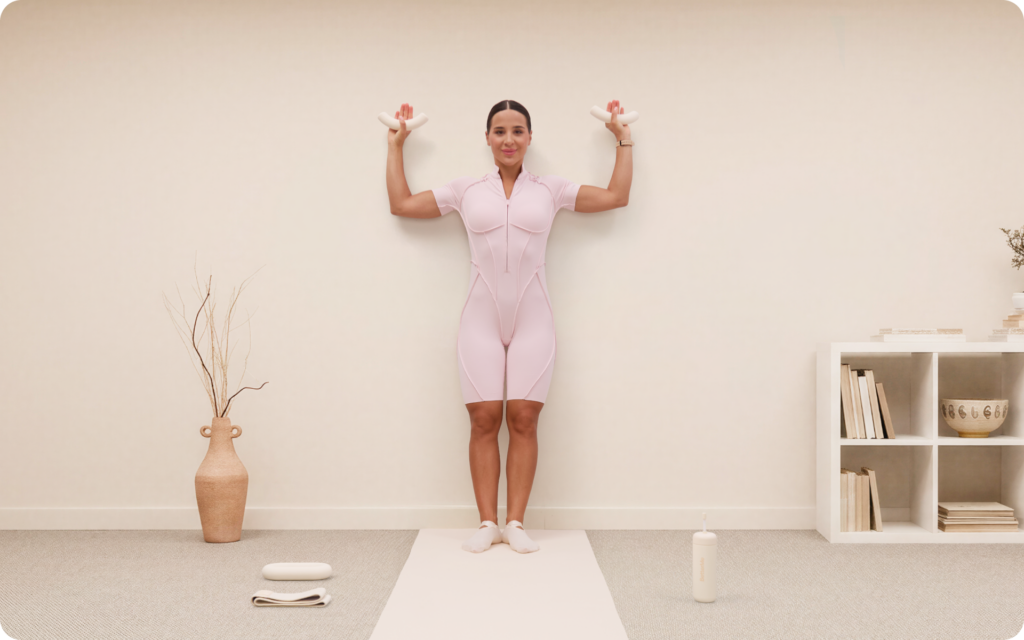 Woman standing against a wall on a yoga mat while holding small ivory dumbbells with her arms bent in a minimal beige workout room.