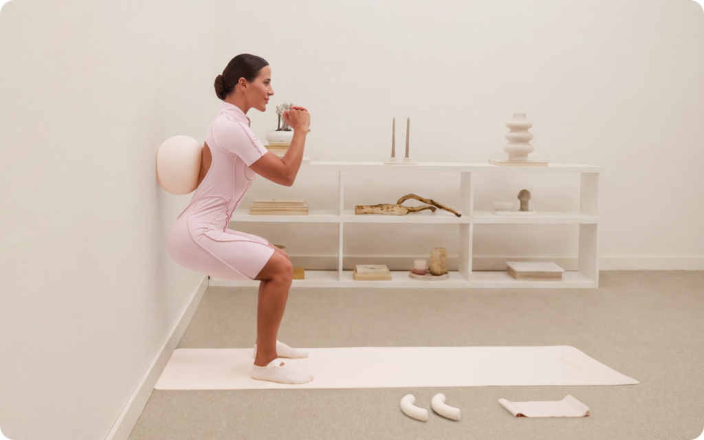 Woman performing a wall squat with a Pilates ball behind her back on a mat in a minimal beige workout room.