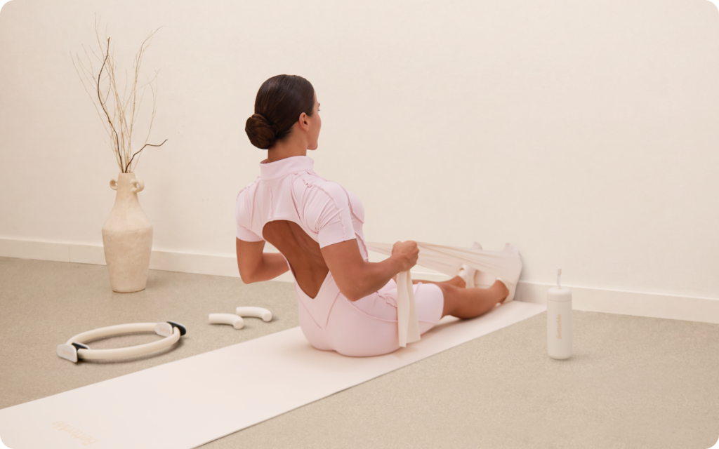 Woman sitting on a mat with her feet against a wall while pulling a resistance band in a minimal beige workout room.