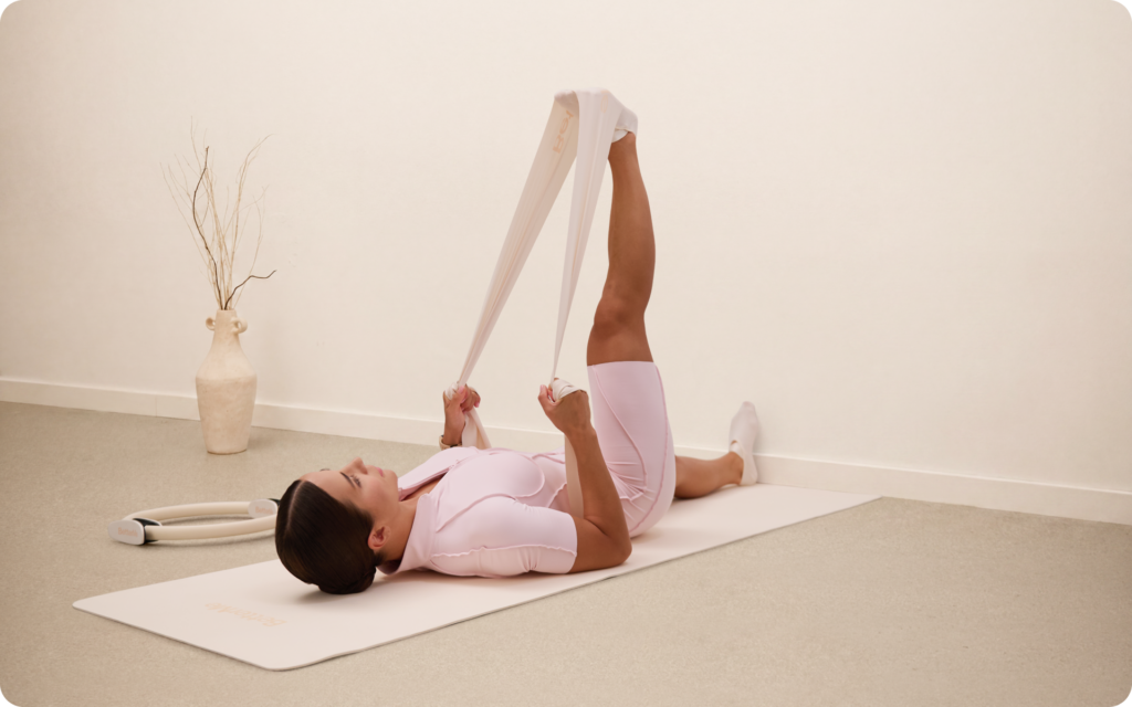 Woman lying on a mat while stretching one leg upward with a resistance band around her foot in a minimal beige workout room.