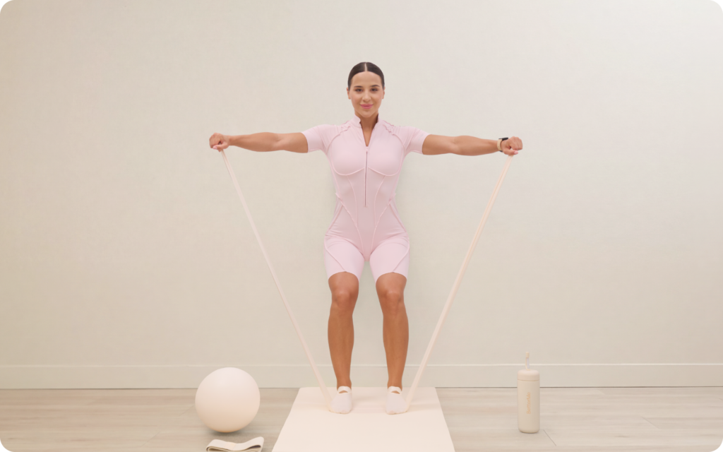 Woman holding a wall squat while stretching a resistance band with both arms on a mat in a minimal beige workout room.