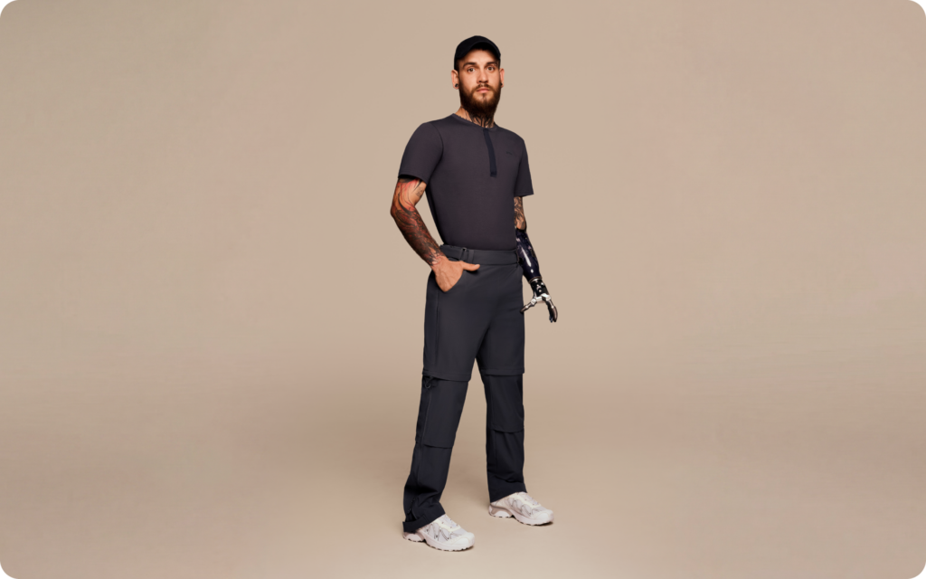 Man with a prosthetic arm wearing adaptive clothing: navy adjustable pants, a dark short-sleeve top, and white sneakers in a minimal studio setting.