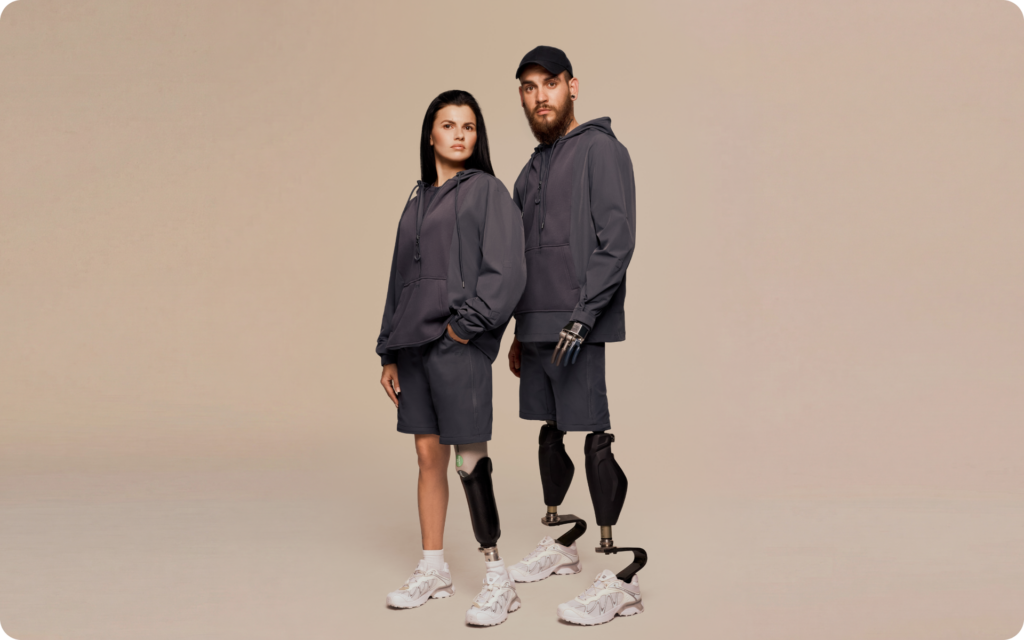 Two models with prosthetic legs wearing adaptive clothing: matching navy hoodies and shorts styled with white sneakers in a minimal studio setting.